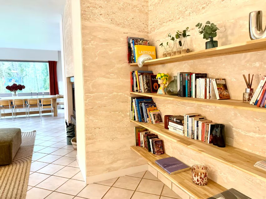 Le mur de pierre claire et ses étagères de livres, avec la pièce de vie en arrière-plan